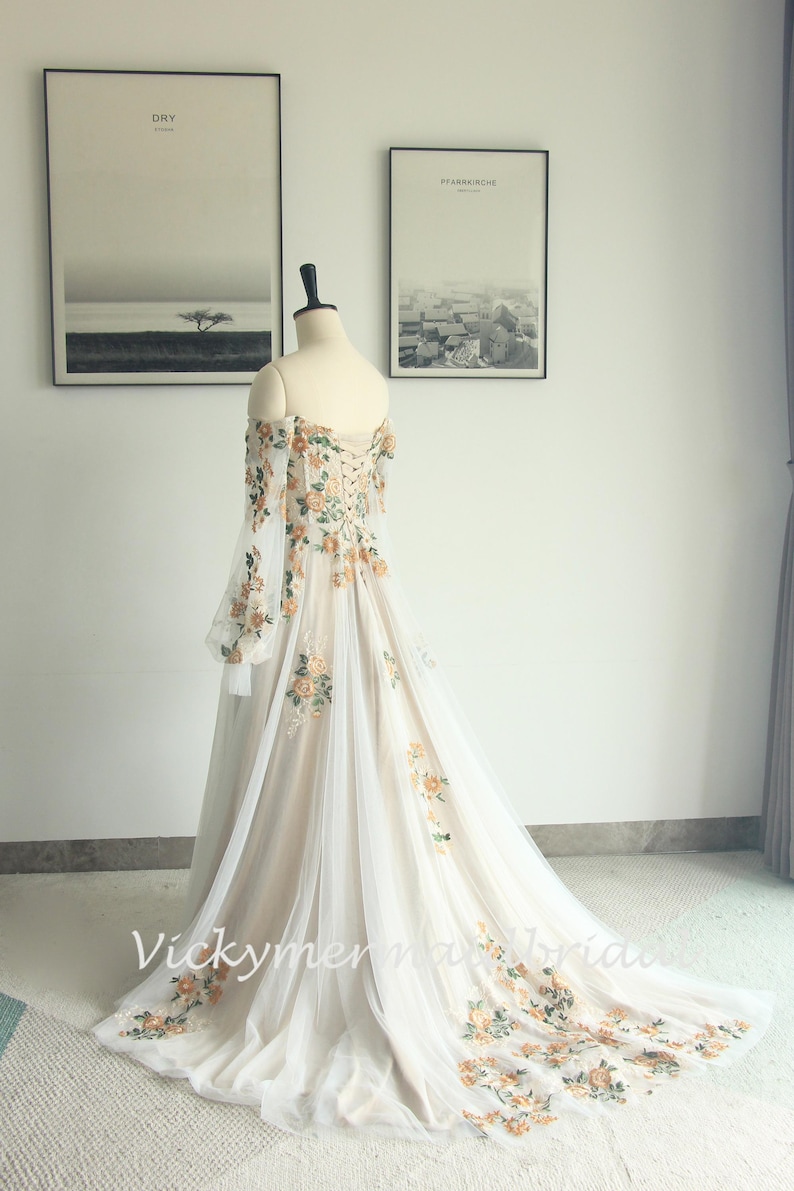 May include: Off-the-shoulder wedding gown with floral embroidery. The ivory dress features long, sheer sleeves and a flowing train. The bodice and train are adorned with orange and green floral accents. The dress is displayed on a mannequin.