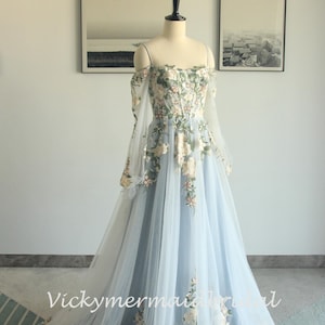 May include: A light blue off-the-shoulder gown with floral embroidery. The dress features long, sheer sleeves and a flowing skirt. The bodice and skirt are adorned with delicate, cream-colored floral patterns. The dress is displayed on a mannequin.