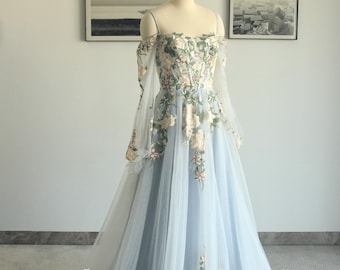 May include: A light blue off-the-shoulder gown with floral embroidery. The dress features long, sheer sleeves and a flowing skirt. The bodice and skirt are adorned with delicate, cream-colored floral patterns. The dress is displayed on a mannequin.