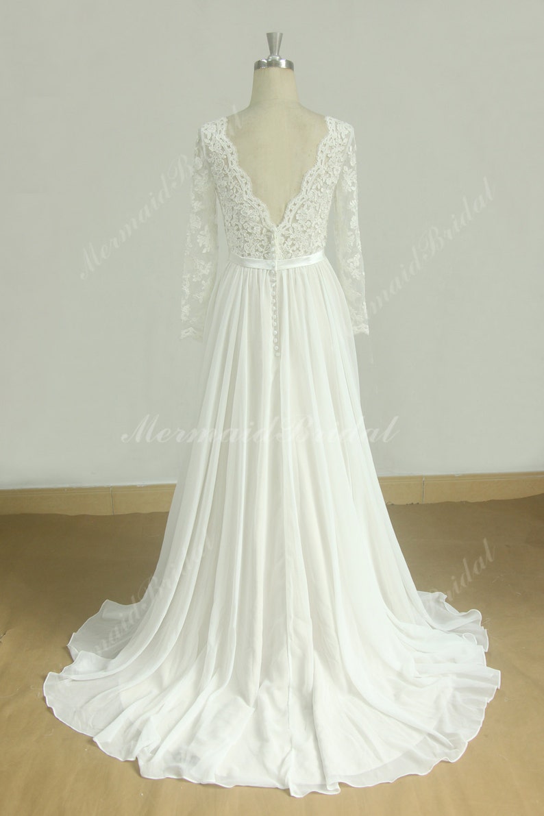 May include: A white chiffon wedding dress with long lace sleeves and a deep V-neckline. The dress has a fitted bodice and a flowing skirt.