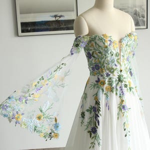 Abito da sposa in chiffon e pizzo a trapezio, stile fiabesco, con ricami colorati e maniche a campana con spalle scoperte e corsetto sulla schiena.