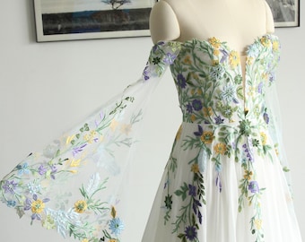 May include: White off-the-shoulder wedding dress with floral embroidery in yellow, purple, and green. The dress features sheer sleeves and a plunging neckline. The text "Vickymermaidbridal" is visible on the dress.