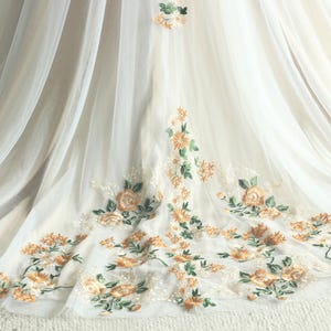 May include: Ivory-colored fabric with delicate floral embroidery in shades of peach, orange, and green. The embroidery features roses and other flowers, creating a romantic and elegant design. The fabric appears to be layered, with a sheer overlay.