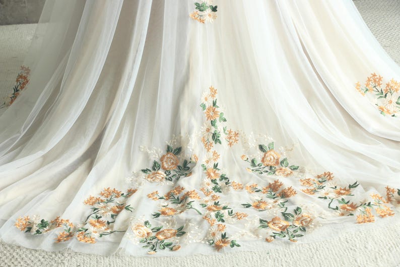 May include: Ivory-colored fabric with delicate floral embroidery in shades of peach, orange, and green. The embroidery features roses and other flowers, creating a romantic and elegant design. The fabric appears to be layered, with a sheer overlay.
