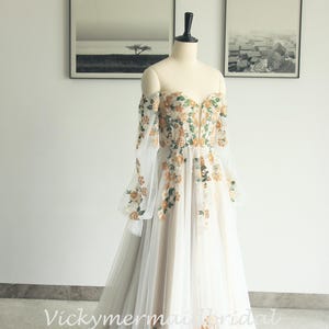 May include: Off-the-shoulder wedding dress with floral embroidery. The ivory gown features long, sheer sleeves and a flowing tulle skirt. The bodice and sleeves are adorned with orange and green floral details. The dress is displayed on a mannequin.