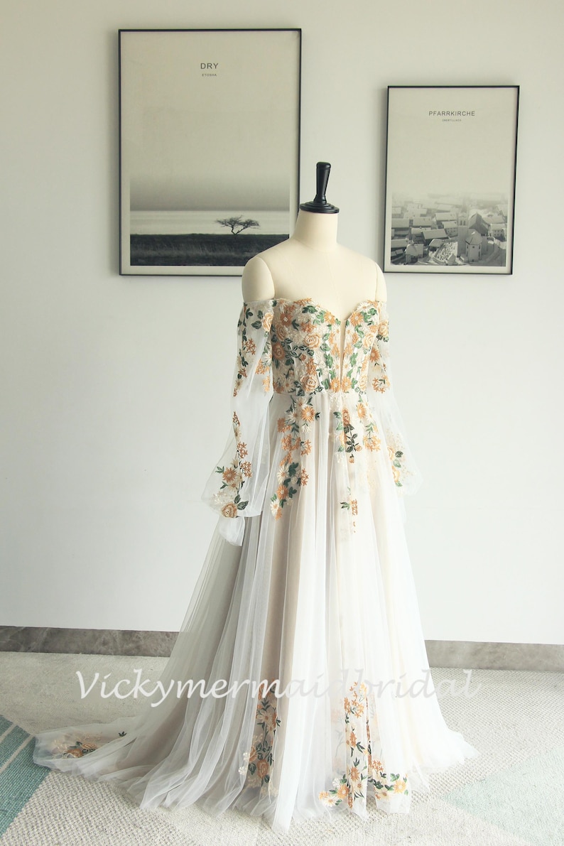 May include: Off-the-shoulder wedding dress with floral embroidery. The ivory gown features long, sheer sleeves and a flowing tulle skirt. The bodice and sleeves are adorned with orange and green floral details. The dress is displayed on a mannequin.