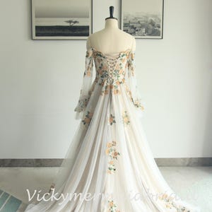 May include: Off-the-shoulder ivory wedding gown with floral embroidery. The long-sleeved dress features a corset back and a flowing tulle skirt, adorned with yellow and green floral accents. The dress is displayed on a mannequin.