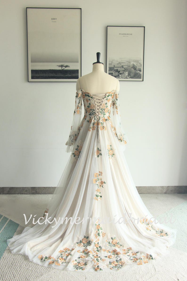 May include: Off-the-shoulder ivory wedding gown with floral embroidery. The long-sleeved dress features a corset back and a flowing tulle skirt, adorned with yellow and green floral accents. The dress is displayed on a mannequin.