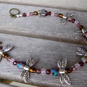 May include: A silver dragonfly charm bracelet with pink, brown, and clear glass beads. The bracelet has a silver clasp and is accented with silver dragonfly charms.