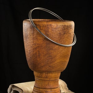 Puede incluir: Un tambor djembé de madera con un acabado de madera natural y un anillo de metal alrededor de la parte superior. El tambor está sentado sobre una alfombra de piel de animal blanca y marrón.