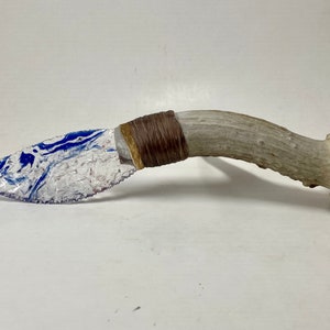 May include: A handmade knife with a blue and white marbled obsidian blade and a brown leather wrapped antler handle.