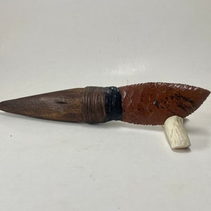 May include: A handcrafted knife with a red and brown flint blade, a wooden handle wrapped in brown leather, and a black accent. The knife is resting on a piece of white antler.