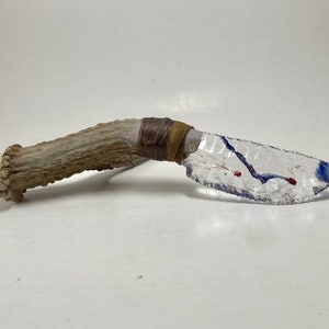 May include: A clear glass knife with blue and red streaks, attached to a brown antler handle with brown leather wrappings.