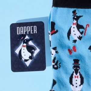 May include: A pair of blue socks with a pattern of penguins wearing top hats and bow ties. The socks are next to a card with the word "Dapper" and a penguin wearing a top hat and bow tie.