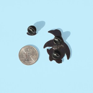 May include: Black enamel pin of a cartoon turtle with a black pin back. The pin is on a light blue background.
