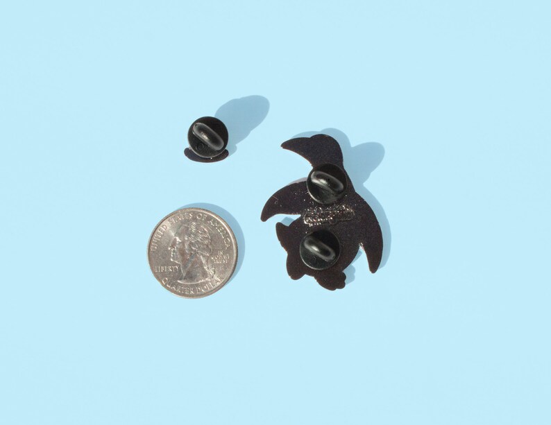 May include: Black enamel pin of a cartoon turtle with a black pin back. The pin is on a light blue background.