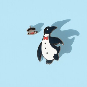 May include: A black and white penguin enamel pin with a red bow tie. The penguin is wearing a tuxedo and has a top hat.