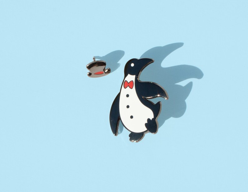 May include: A black and white penguin enamel pin with a red bow tie. The penguin is wearing a tuxedo and has a top hat.