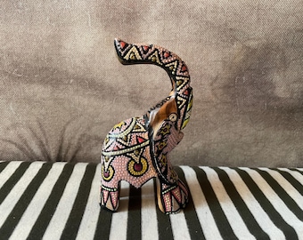 May include: A decorative wooden elephant figurine with an upturned trunk. The elephant is painted with pink, yellow, and black geometric patterns. The figurine is displayed on a black and white striped surface.