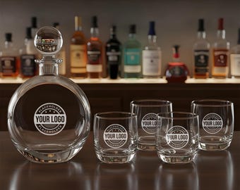 May include: A clear glass decanter with a round body and a spherical stopper, accompanied by four matching lowball glasses. Each glass and the decanter feature a circular logo that says "YOUR LOGO". The set is displayed on a dark wooden surface, with bottles in the background.