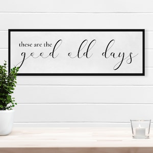 May include: A black framed sign with white background and the text "these are the good old days" written in black cursive font.