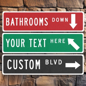 May include: Three black and white street signs with white text and arrows. The top sign is red and says "Bathrooms Down" with a white down arrow. The middle sign is green and says "Your Text Here" with a white right arrow. The bottom sign is black and says "Custom Blvd" with a white right arrow.