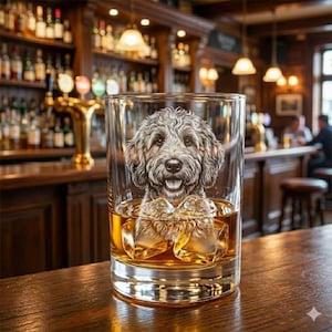May include: A clear, square-shaped rocks glass with a detailed etched image of a dog's face. The glass is filled with amber-colored liquid and ice cubes. The glass sits on a wooden bar in a pub setting.