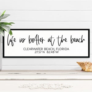 May include: Black and white sign with the text "Life is better at the beach" and the location "Clearwater Beach, Florida 27.57 N 82.48 W".