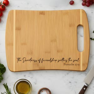 May include: A rectangular bamboo cutting board with a handle. The board features the engraved text "The Sweetness of friendship soothes the soul" and "Proverbs 27:9". The board is surrounded by fresh ingredients.