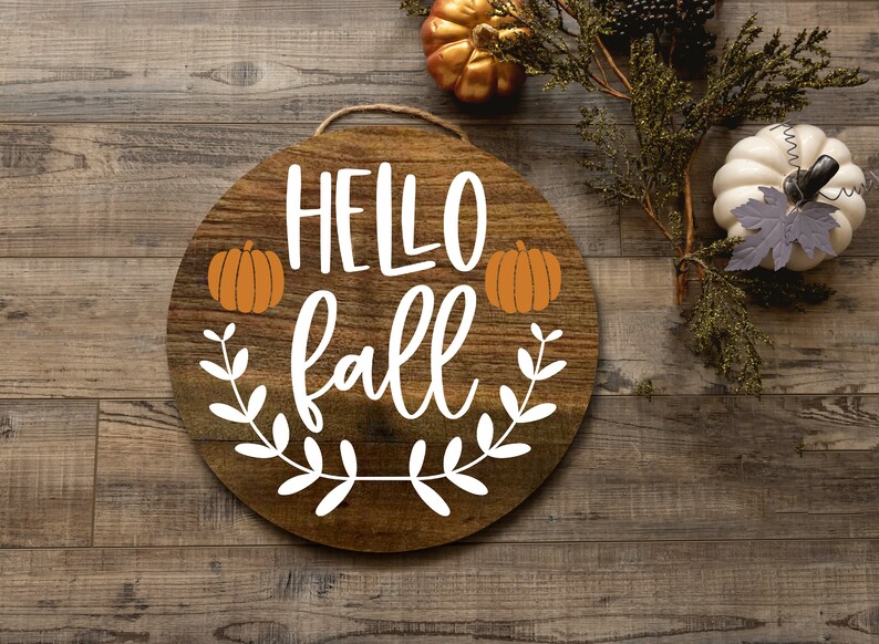 Pode incluir: Um sinal redondo de madeira com o texto "HELLO fall" em letras brancas. O sinal &eacute; decorado com folhas brancas e duas ab&oacute;boras laranja.