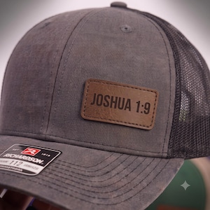 May include: A charcoal gray trucker hat featuring a brown rectangular faux leather patch centered on the front panel. The patch is engraved with the text JOSHUA 1:9 in a bold sans-serif font. The hat includes a black mesh back and a sticker.