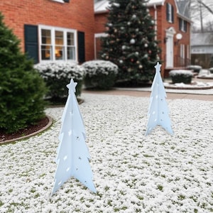 May include: Two white wooden Christmas trees with star toppers stand in a snowy yard. The trees are decorated with cut-out stars.