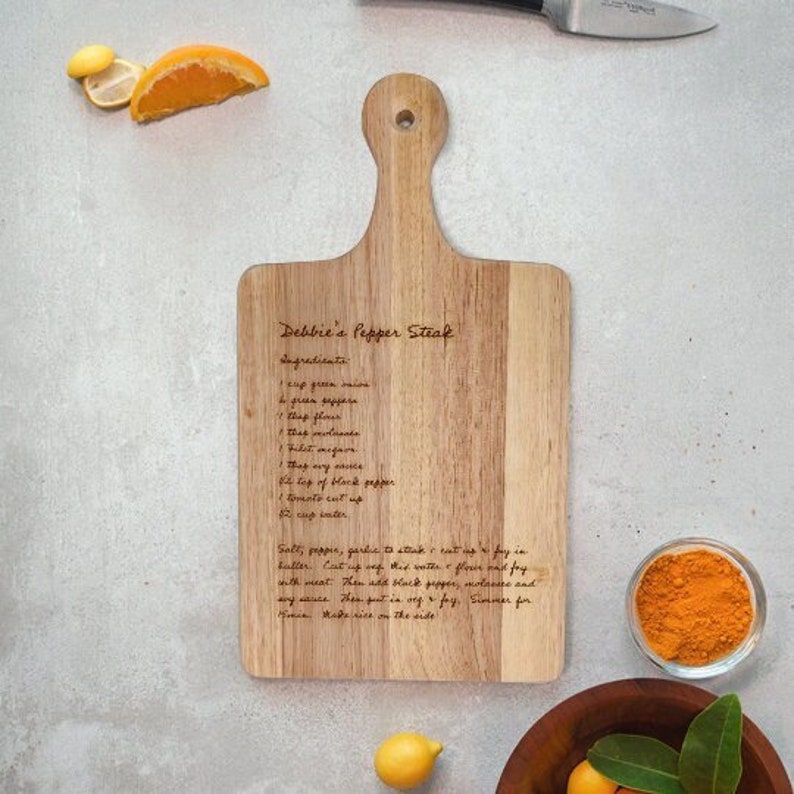 Puede incluir: Una tabla de cortar de madera con un asa, con una receta de "Debbie's Pepper Steak" grabada en su superficie. La tabla est&aacute; rodeada de ingredientes como limones, rodajas de naranja y un peque&ntilde;o cuenco de polvo naranja. Un cuchillo es visible en el fondo.