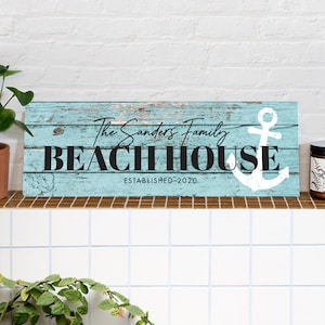 Op de afbeelding: Een lichtblauw houten bord met een wit anker en de tekst "The Sanders Family Beach House Established 2020".