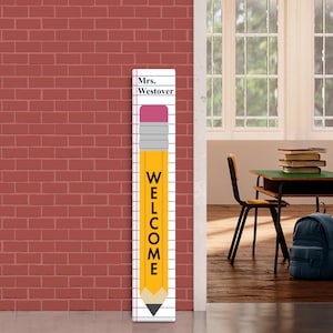 Op de afbeelding: Een lang, rechthoekig welkomstbord ontworpen als een gele potlood met een roze gum. Het woord "WELCOME" staat verticaal op het potlood gedrukt. Het bord bevat ook de naam "Mrs. Westover". Het bord staat voor een rode bakstenen muur.