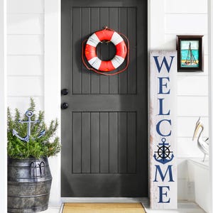 May include: A white wooden sign with the word "WELCOME" in blue letters. The sign has a nautical theme with a blue anchor and a ship's wheel. The sign is leaning against a white door with a red and white life preserver hanging on it.
