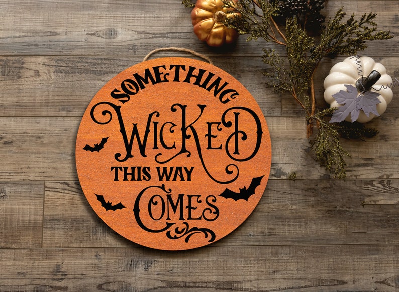 May include: A round wooden sign with the text "Something Wicked This Way Comes" in black lettering. The sign is decorated with bats and swirls.