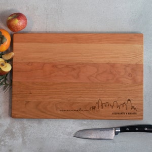 May include: A rectangular wooden cutting board with a cityscape design and the word "cincinnati" engraved on it. The names "Stephanie & Marcus" are also engraved. A knife and fruit are next to the board.