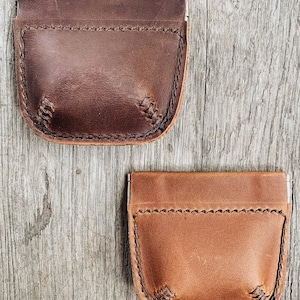 May include: Two leather coin purses with a flap closure and a stitched design. The purse on top is brown and the purse on the bottom is tan.