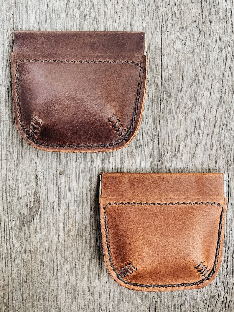 May include: Two leather coin purses with a flap closure and a stitched design. The purse on top is brown and the purse on the bottom is tan.