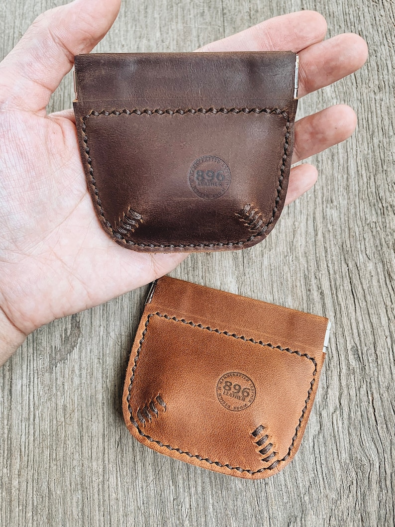 May include: Two leather coin purses with a snap closure. The purses are brown and tan and have a stitched baseball design. The purses have a circular logo that says "896 Leather".
