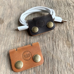 May include: Two leather cord organizers with brass accents. The top organizer is brown leather and the bottom organizer is tan leather. Both organizers have the text "896 Leather" embossed on them.