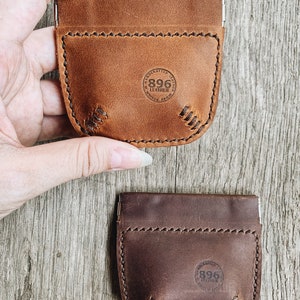 May include: Two leather coin purses with a snap closure. The purse on the left is brown and has a stitched design. The purse on the right is dark brown and has a stitched design. Both purses have a circular logo with the text "896 Leather" and "Handcrafted Goods".