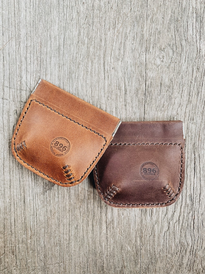 May include: Two leather coin purses with a stitched edge and a circular logo that says "896 Leather". The purse on the left is brown and the purse on the right is dark brown.