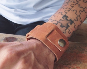 May include: A brown leather cuff bracelet with a metal button closure. The bracelet is worn on a person's wrist.