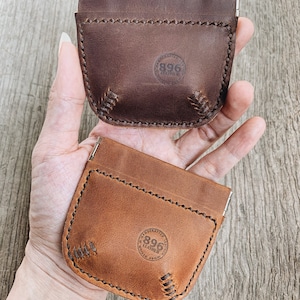 May include: Two leather coin purses with a snap closure. The purses are brown and tan and have a stitched design. The purses have a circular logo with the text "896 Leather" on them.