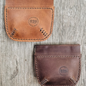 May include: Two leather coin purses with a snap closure. The top purse is brown and the bottom purse is dark brown. Both purses have a stitched design and a circular logo that says "896 Leather" in the center.