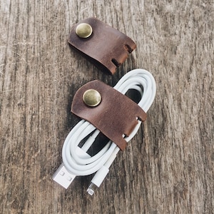 May include: Two brown leather cord organizers with brass buttons. One organizer is holding a white charging cable.