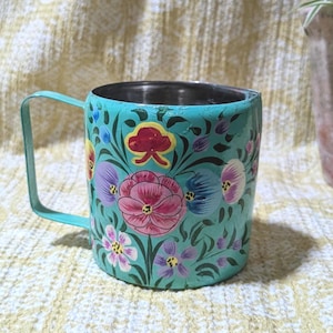 May include: A teal metal mug with a floral design. The mug has a stainless steel interior and a teal handle. The exterior is decorated with hand-painted flowers in pink, purple, yellow, and red. The mug is a decorative item.