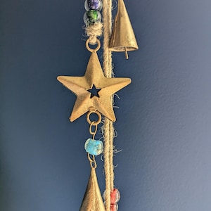 May include: A rustic wind chime with a star and bell design. The chime features a bronze star, two bronze bells, and colourful glass beads strung on natural twine. The background is a solid dark blue.